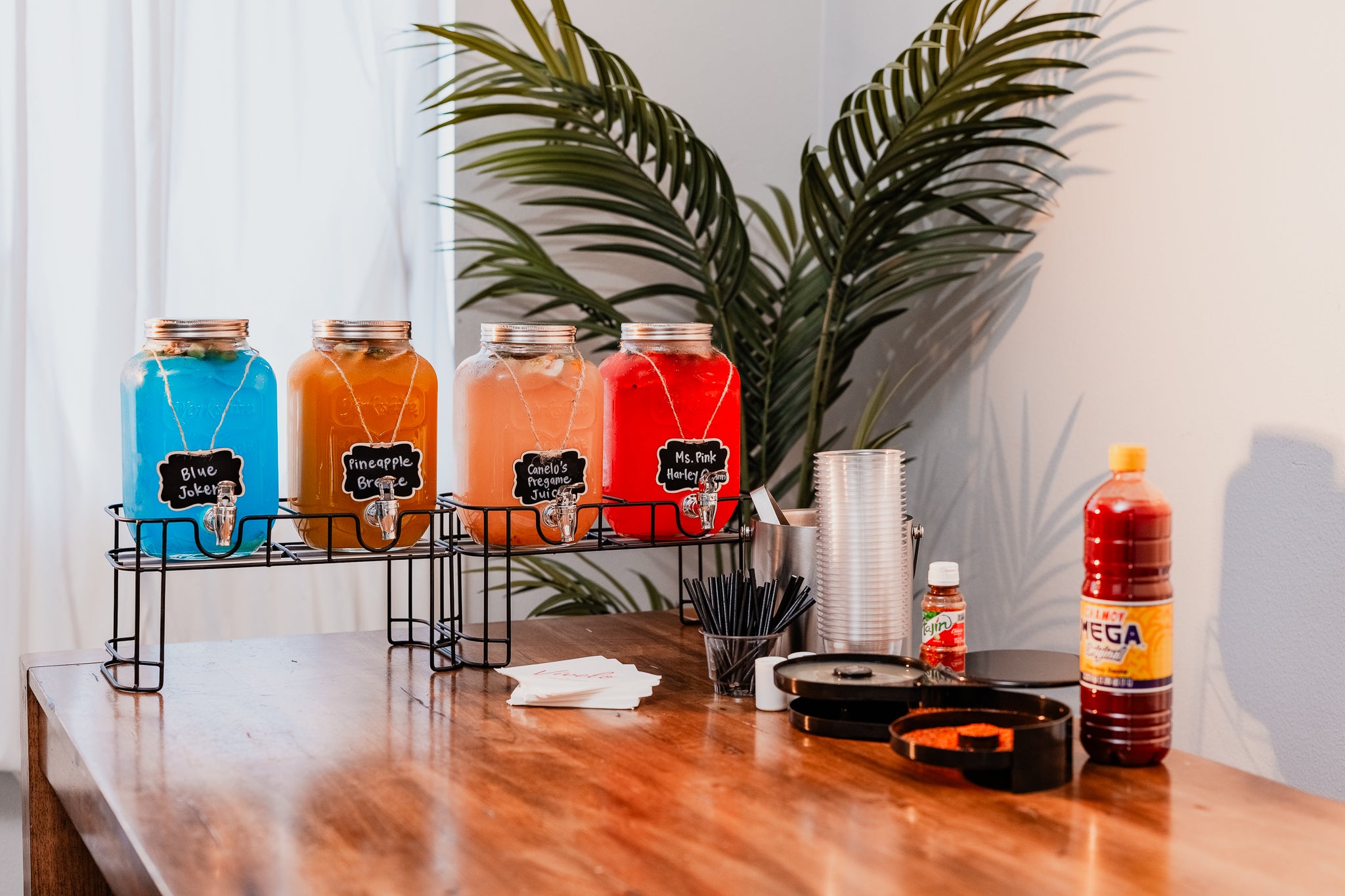 Assorted cocktail drinks in glass dispensers on a wooden table with a plant in the background