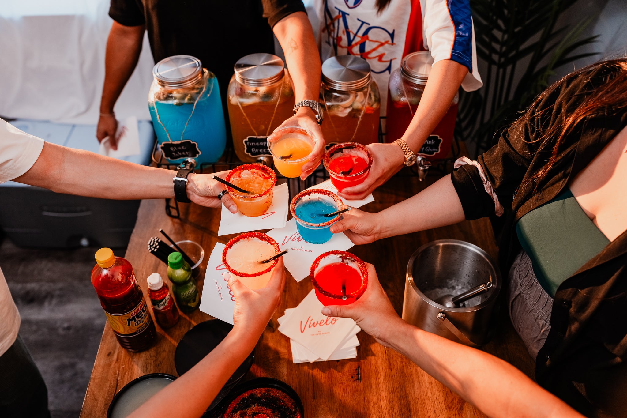People holding colorful cocktail drinks at a table with jars and bottles.