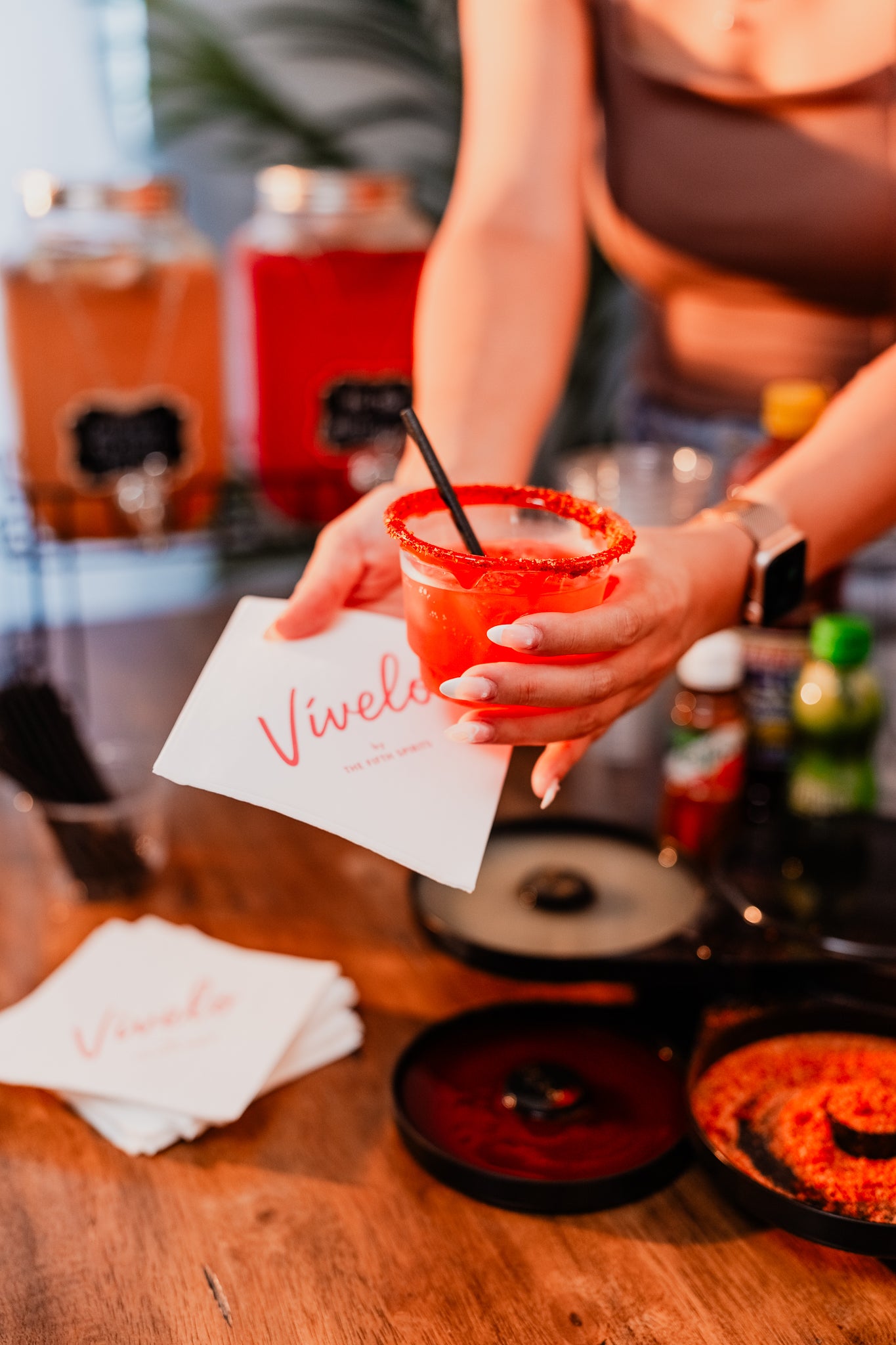 Person holding a cocktail cup with a straw and a 'Vivelo' napkin on the wooden table with cocktail jar dispensers in the back