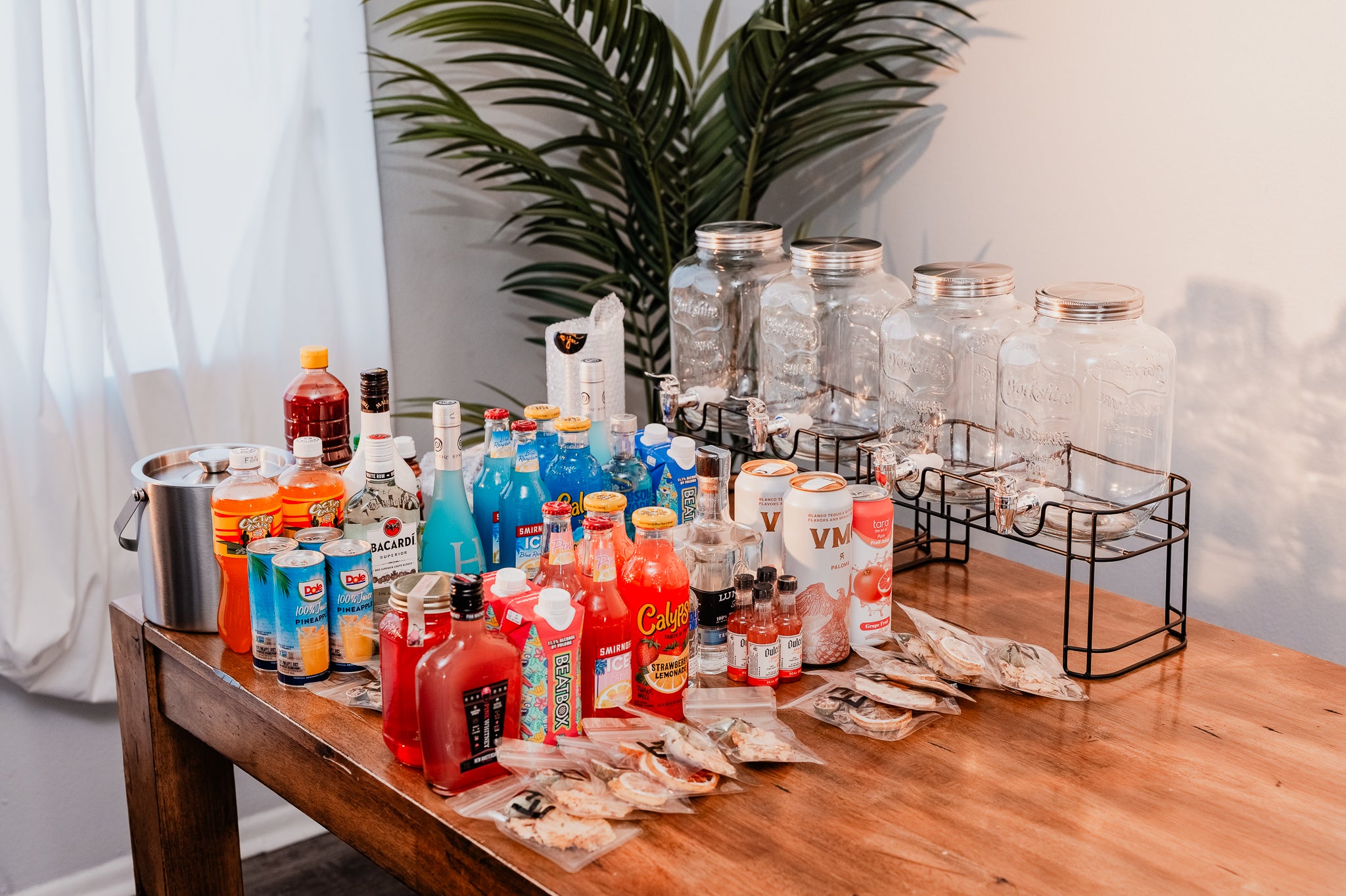 Table with various bottles of alcohol and mixers in a room with plants and a curtain.